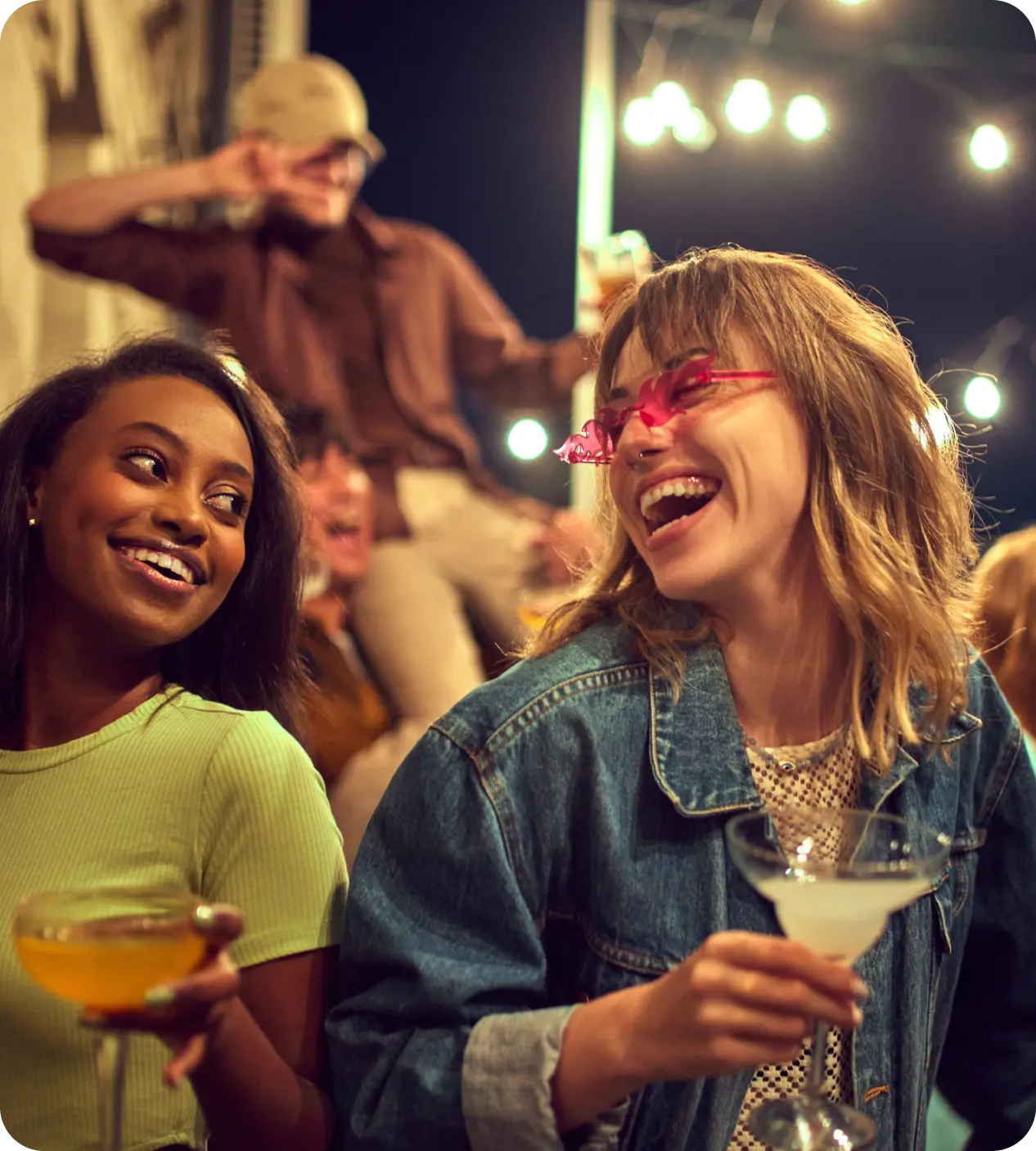 Two women enjoying drinks and laughter together at a lively social gathering.