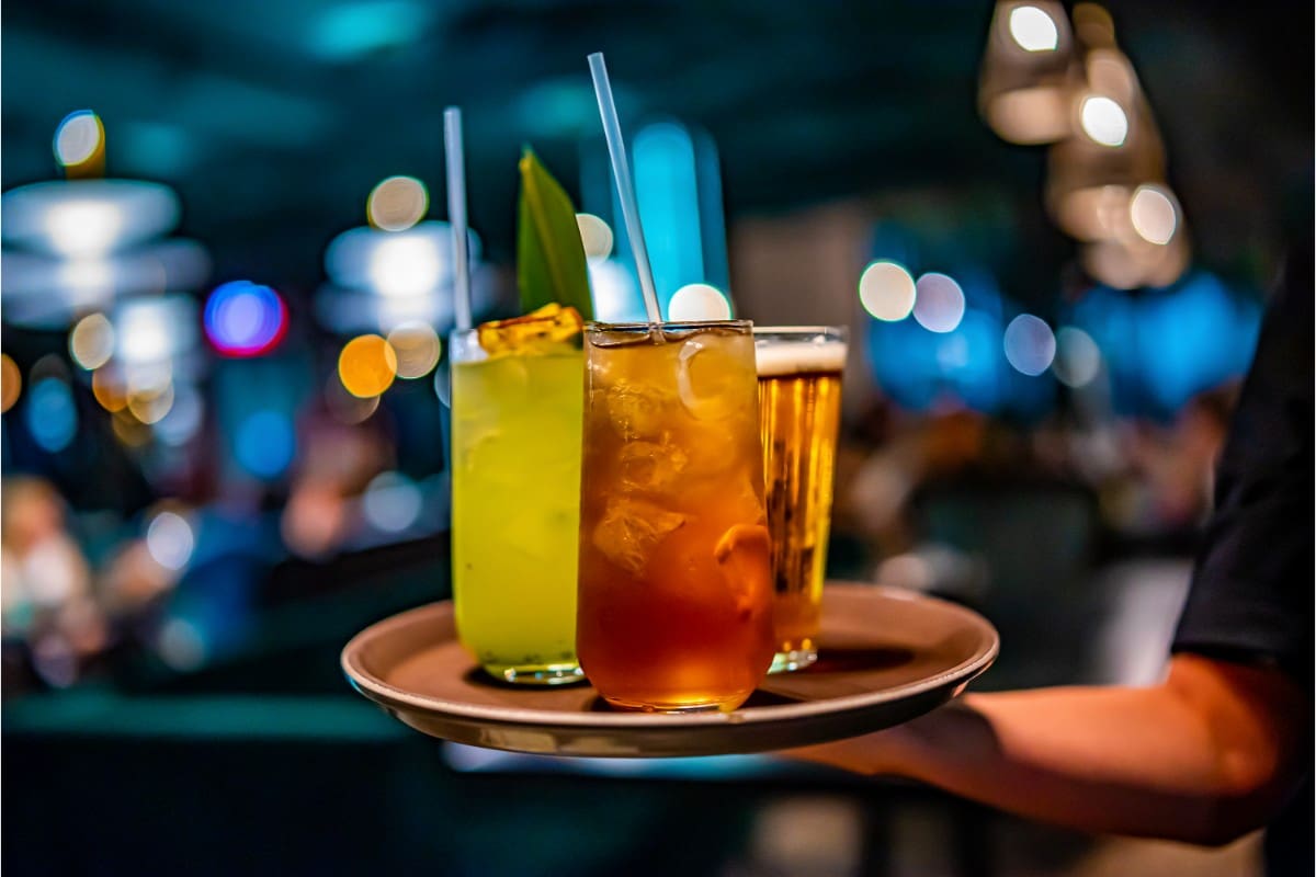 Three colorful cocktails in glasses on a tray with straws and garnishes.