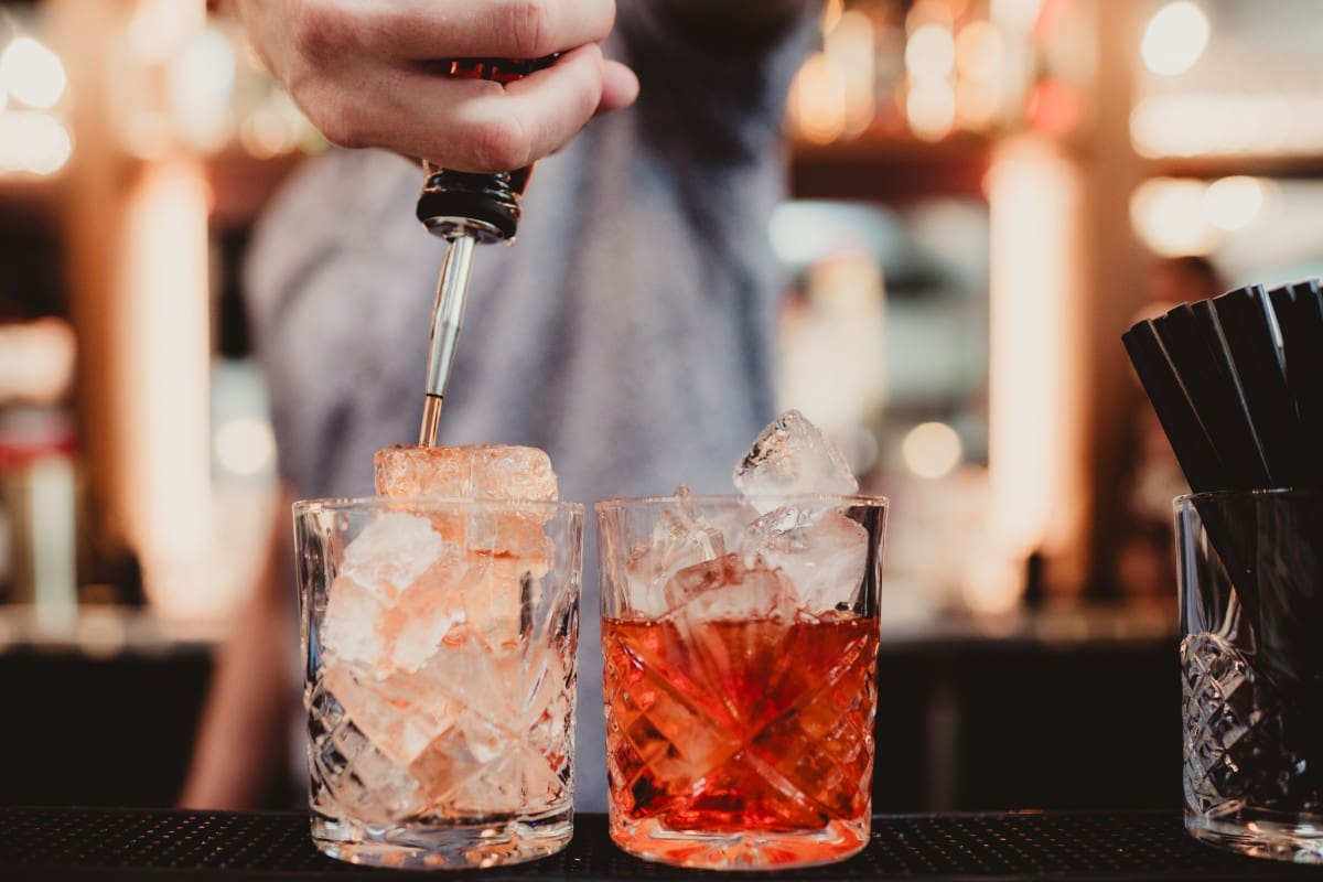 Bartender pouring a drink over ice in two glasses at a bar.