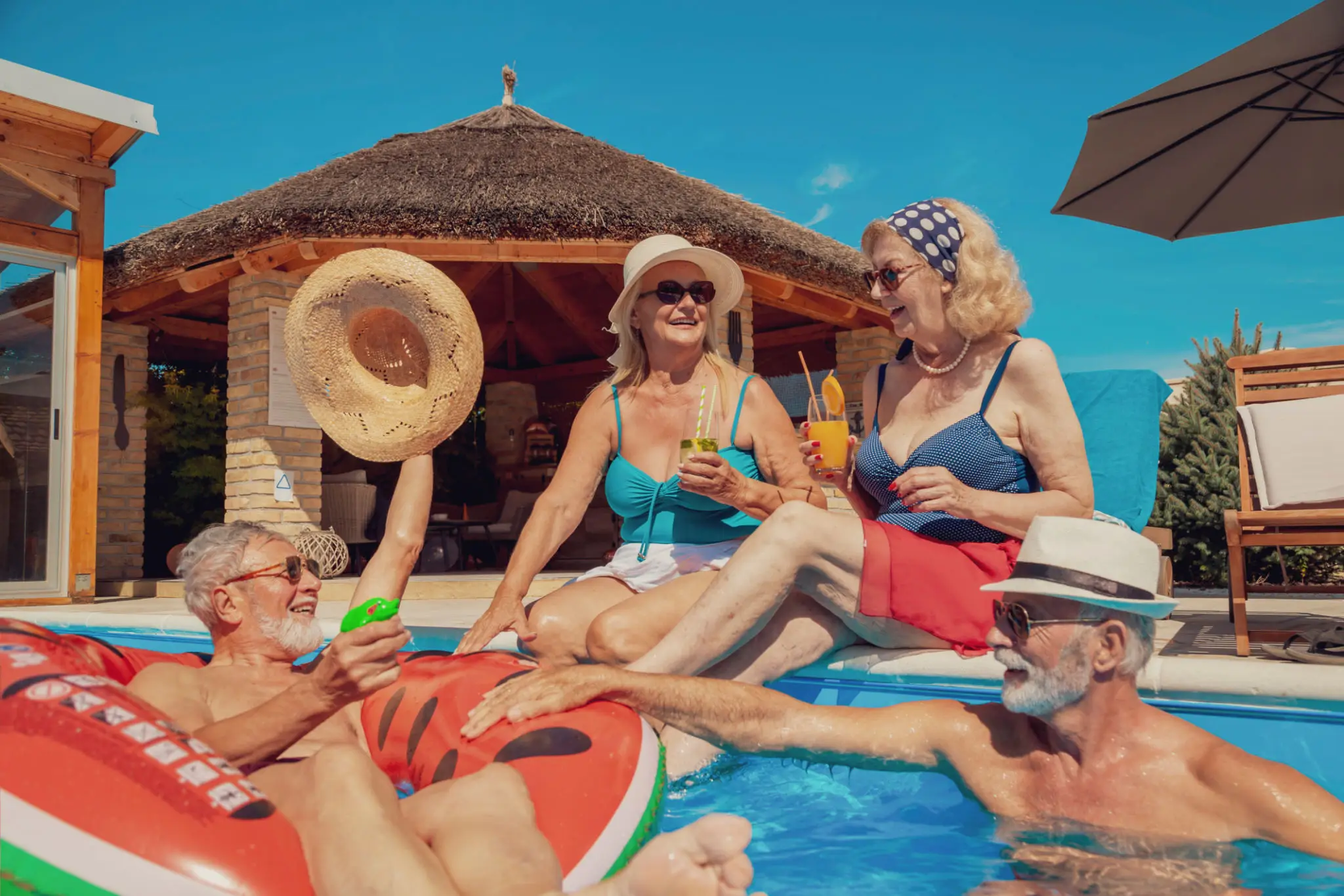 Four seniors enjoying a sunny pool day with drinks and laughter.